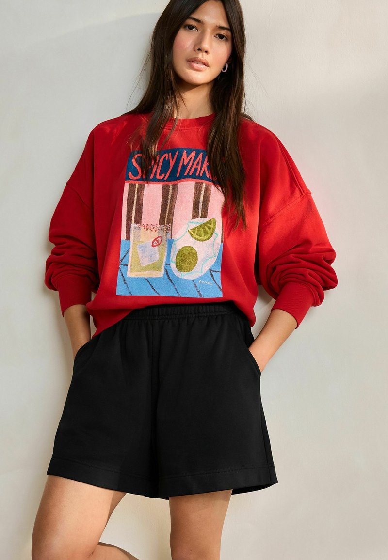 Rød sweatshirt med et farverigt grafisk motiv med drikkevarer, kombineret med sorte shorts. Begge dele er afslappede og har en løs pasform.