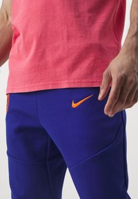Nike Sportbekleidung zeigt ein korallenfarbenes T-Shirt und blaue Jogginghosen mit einem orangefarbenen Swoosh-Logo, das eine glatte Textur und eine sportliche Passform aufweist.