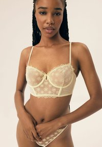 Jasnożółty wiskozowy biustonosz z koronkowymi kwiatowymi wzorami, z przezroczystymi miseczkami, wsparciem w postaci fiszbiny oraz delikatnym falbaniastym wykończeniem na krawędziach. Modelka pozuje z dłońmi złożonymi.