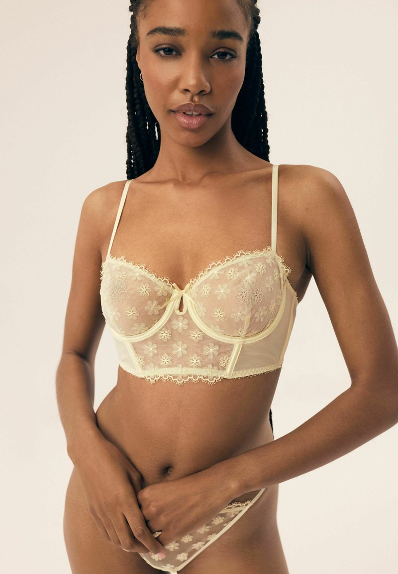 Jasnożółty wiskozowy biustonosz z koronkowymi kwiatowymi wzorami, z przezroczystymi miseczkami, wsparciem w postaci fiszbiny oraz delikatnym falbaniastym wykończeniem na krawędziach. Modelka pozuje z dłońmi złożonymi.