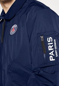 Laivaston tuulenpitävä takki, jossa on kauluksellinen kaula, vetoketjullinen tasku vasemmassa hihassa ja brodeerattu Paris Saint-Germain -logo rinnassa.