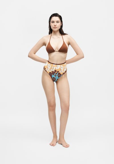 Γυναίκα όρθια ξυπόλητη με τα χέρια στη μέση, ντυμένη με καφέ τοπ μαγιό halter και ψηλόμεσο μπικίνι με λουλούδια σε λευκό φόντο.