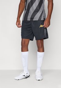 Svarte sportsshorts med gulllogo, kombinert med hvite knelange sokker og hvite fotballsko, iført en grå og svartstripet trøye.