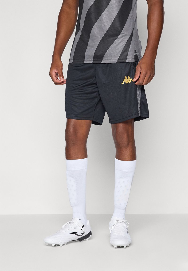 Svarte sportsshorts med gulllogo, kombinert med hvite knelange sokker og hvite fotballsko, iført en grå og svartstripet trøye.