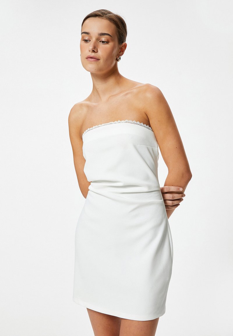 Mini vestido blanco sin tirantes con adornos de encaje en el escote, fabricado en una tela suave, que presenta una silueta ajustada y un diseño minimalista.