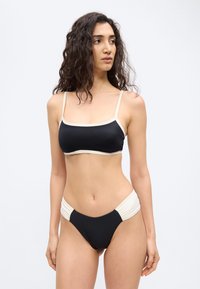 Γυναίκα με σγουρά σκούρα μαλλιά, που φοράει μαγιό μπικίνι ασπρόμαυρο, στέκεται και κοιτάζει προς τα αριστερά της, μπροστά σε ένα απλό φωτεινό φόντο.