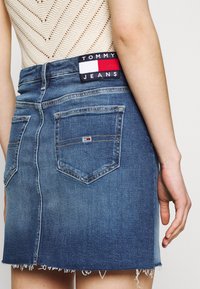 Denim minikjol i blått med fransar i nederkant, med två bakfickor och ett märkespatch vid midjan med röd, vit och blåa inslag.