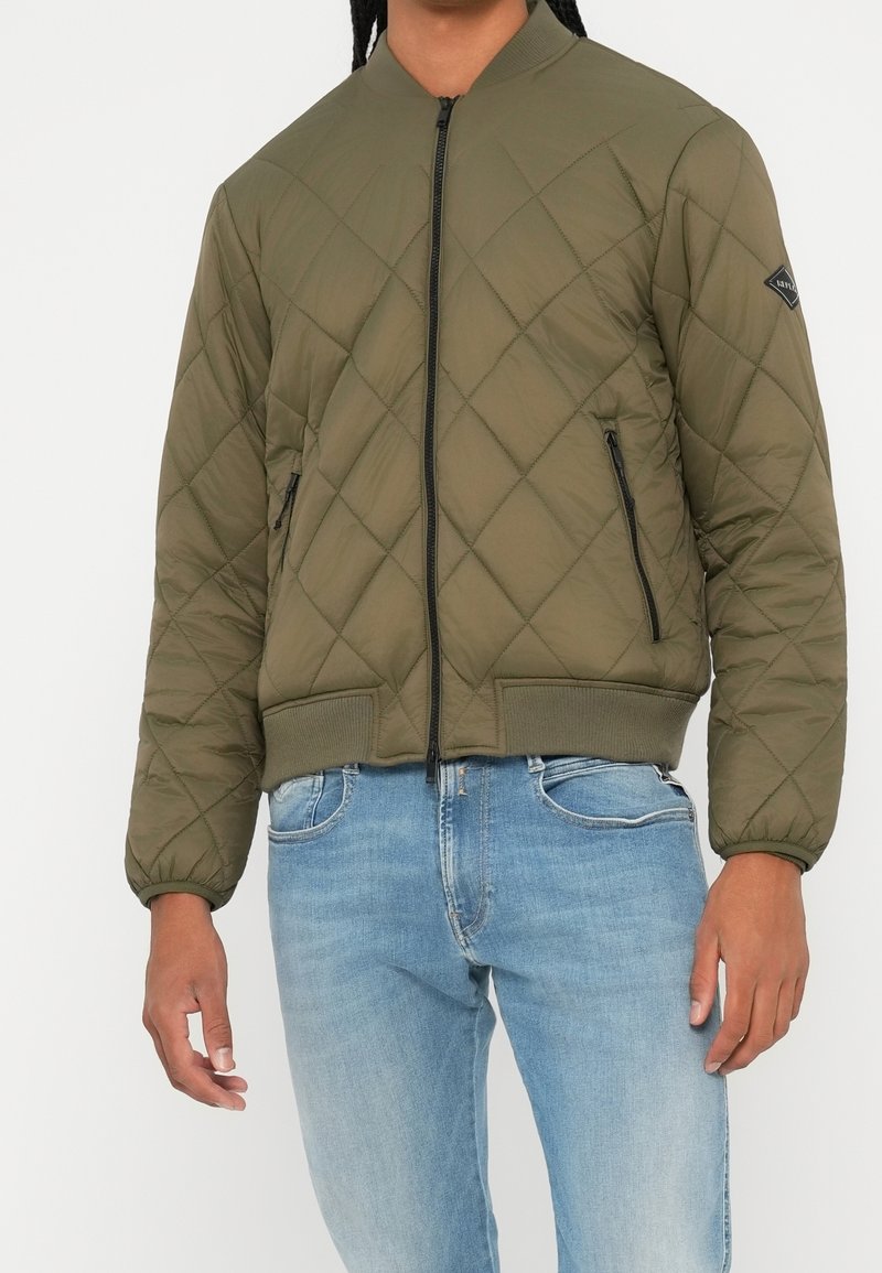 Homme portant une veste bomber matelassée vert olive avec poches zippées et un jean bleu clair sur un fond uni.