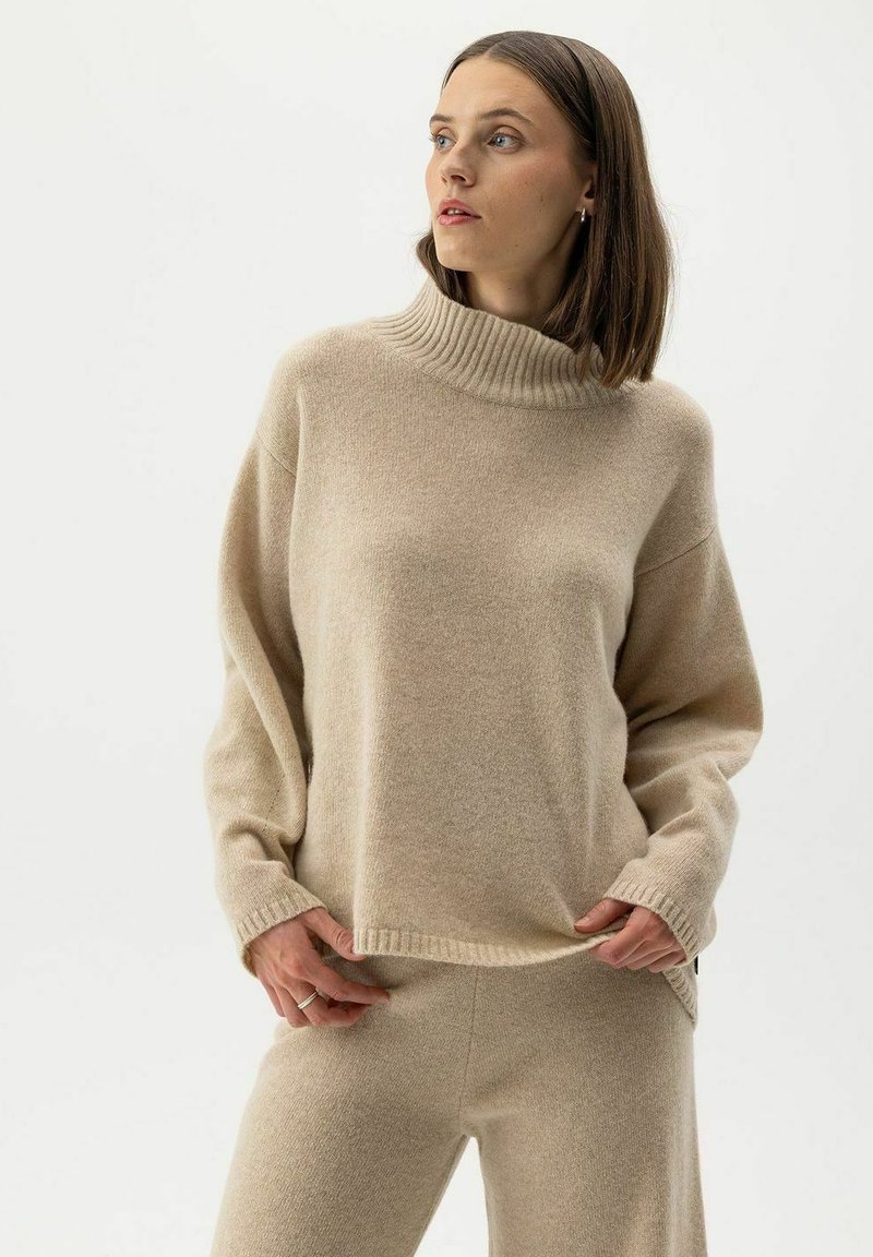Frau, die einen beigen Strick-Turtleneck-Pullover und passende Hosen trägt, steht mit den Händen, die den Saum des Pullovers halten, und schaut zur Seite.
