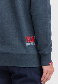 Sudadera gris oscuro con puños y dobladillo acanalados. Presenta texto bordado en rojo, "!SLD© Desde 1993," en la parte inferior y en la manga. Textura suave.