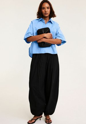 Frau trägt ein locker sitzendes hellblaues Hemd und weite schwarze Hosen, hält eine schwarze Clutch und steht in schwarzen Sandalen mit Absatz vor einfarbigem Hintergrund.