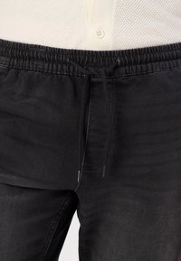 Pantalons noirs avec une taille élastique et un cordon de serrage. Le tissu est un denim texturé, présentant une apparence légèrement décolorée.