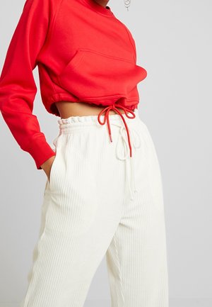 Osoba v červené krátké mikině se stahovací šňůrkou a vysokým pasem bílých žebrovaných kalhotách s kapsami, stojící na jednotném pozadí.