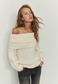 Ekrasti pleteni pulover z rebrasto teksturo, izrezom čez ramena in širokimi rokavi, kombiniran s temnosivimi kavbojkami.