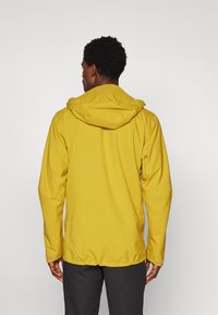 Gul vandtæt jakke med hætte, der har justerbare manchetter og en glat tekstur. Bagfra ses den slanke, moderne design.