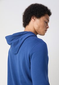Felpa con cappuccio blu dal tocco morbido, collo curvo e cordini regolabili, che mostra il retro e il profilo laterale della persona.