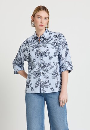 Frau trägt blau gestreiftes Hemd mit schwarzem Blumendruck und halblangen Ärmeln, kombiniert mit blauer Jeans, steht mit einer Hand in der Hüfte.