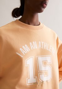 Brzoskwiniowy sweatshirt z prążkowanym okrągłym dekoltem, z napisem "JESTEM SPORTOWCEM" oraz numerem "15" nadrukowanym na biało. Miękka, teksturowana tkanina.