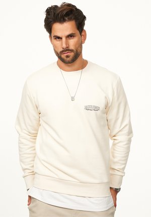 Helles beiges Sweatshirt mit Rundhalsausschnitt, gerippten Bündchen und Saum. Verfügt über ein kleines Logo auf der linken Brust und ist über einem weißen Shirt getragen.