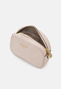 Bolso cruzado de cuero rosa claro con forma redondeada, detalles en dorado y cierre de cremallera. El interior cuenta con un forro con el logo y acentos en color marrón.
