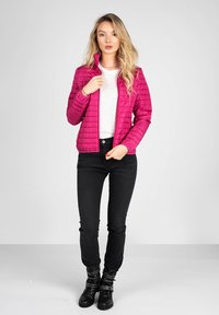 Giacca trapuntata rosa brillante con colletto alto e zip, abbinata a una maglietta grigia chiara, jeans skinny neri e stivaletti neri con accessori.