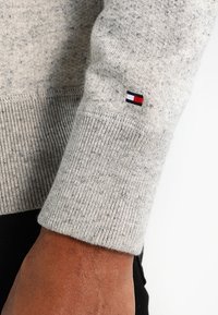 Szary sweter o teksturowanej powierzchni z prążkowanymi mankietami. Posiada małą naszywkę z logo w kolorach czerwonym, białym i niebieskim na lewym rękawie.