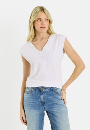 Femme blonde portant un haut blanc sans manches à col en V avec un détail texturé, associée à un jean bleu, debout avec la main sur la hanche devant un fond blanc.