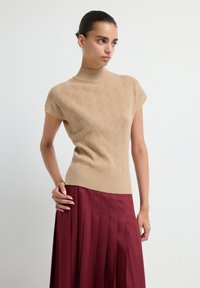 Top de punto beige con cuello alto y mangas cortas, que presenta un patrón de hojas; combinado con una falda burgundy con pliegues.