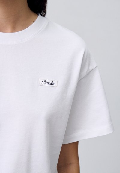 T-shirt bianco in cotone con collo rotondo e maniche corte, con un piccolo etichetta ricamata che dice "Claudie" attaccata sul petto.