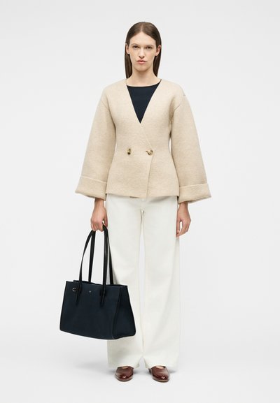 Γυναίκα με μπεζ σακάκι με διπλό κούμπωμα, άσπρο παντελόνι φαρδιάς γραμμής, καφέ παπούτσια, κρατώντας μια μεγάλη μαύρη τσάντα χειρός σε απλό φόντο.