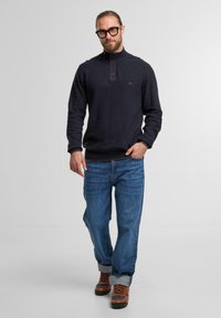 Marineblauer Pullover mit hohem Kragen, geknöpftem Hals und strukturiertem Stoff. Kombiniert mit hellblauen Jeans und braunen Wanderstiefeln.