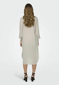 Donna con lunghi capelli mossi che indossa un vestito midi grigio chiaro con maniche arrotolate e sandali neri con tacco alto, in piedi di spalle.