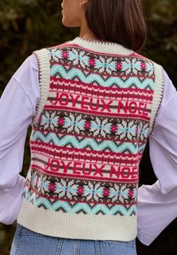 Knit sweater vest in cream, brown, and pink with festive patterns; features text "JOY EUX NOEL," and contrasting ribbed hem.