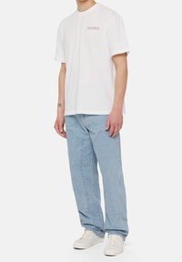 Vit bomullströja med en liten beige "Dickies"-logotyp, kombinerad med ljusblå denimjeans och beige sneakers. Avslappnad passform och stil.