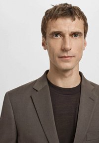 Braunes kariertes Sakko mit Reverskragen über einem dunklen T-Shirt. Der Stoff ist strukturiert mit einem dezenten Muster, und die Passform ist maßgeschneidert.