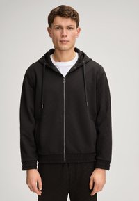 Felpa nera con zip in tessuto testurizzato e cappuccio. Presenta una zip frontale, polsini a coste e una vestibilità comoda. Indossata sopra una maglietta bianca.