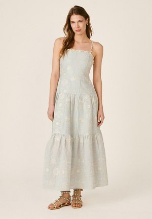 Mujer con vestido maxi sin mangas de color azul claro con bordado floral, dobladillo con volantes y sandalias planas beige con adornos de cuentas.