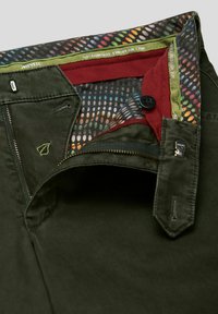 Donkergroene olijfgroene broek met een gestructureerde afwerking, voorzien van een kleurrijke, gestippelde binnenvoering, ritssluiting en contrasterende accenten in rood en groen.