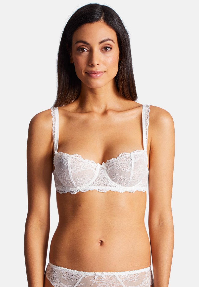 Soutien-gorge en dentelle blanche avec armatures, motif floral complexe, bords festonnés, fines bretelles ajustables et un petit nœud décoratif au centre.