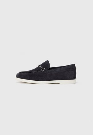 Mocassino in suede grigio scuro con suola in gomma bianca, caratterizzato da un design cucito e da un piccolo accento metallico sulla parte superiore. Stile classico slip-on.