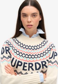 Suéter de punto con cuello redondo, base color crema y patrones geométricos en azul, rosa y azul marino. Sobre una camisa de cuello azul claro.