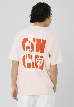 Homme aux cheveux noirs bouclés portant un t-shirt rose pâle avec un texte rouge et des images blanches de bouteille et verre à vin dans le dos.