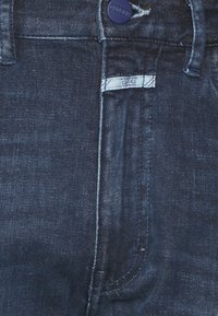 Tmavěmodré džíny z denimu s viditelným knoflíkem a malou bílou značkovou etiketou našitou poblíž oblasti zipu.