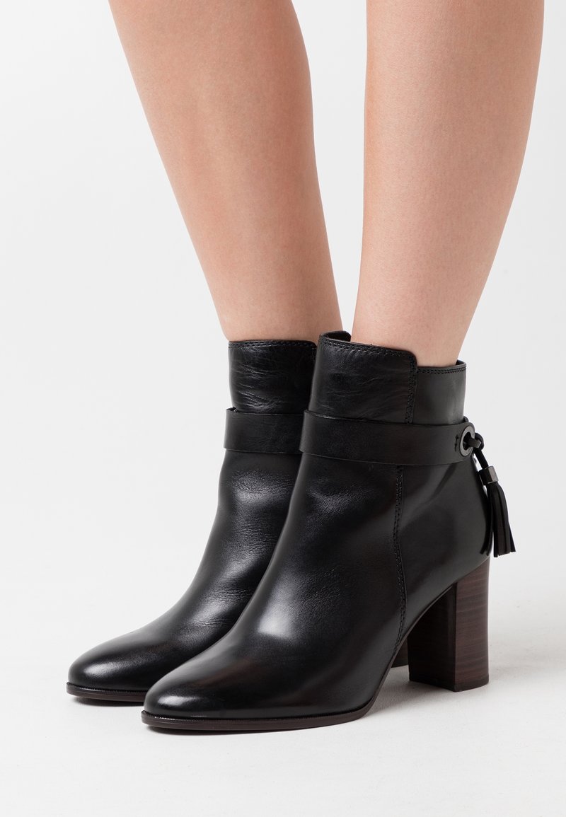 Bottines en cuir noir à talons blocs, bouts pointus et pampilles décoratives portées sur des jambes nues sur un fond blanc uni.