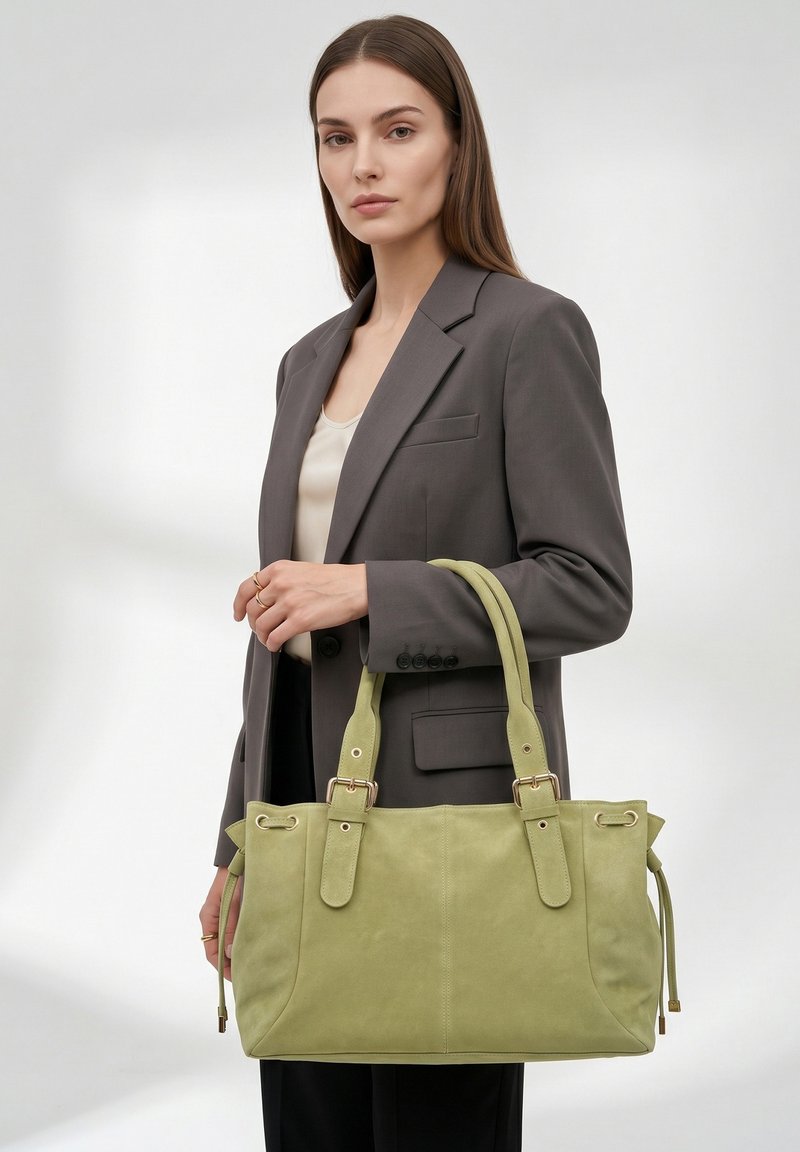 Femme aux longs cheveux bruns portant un blazer gris et un top crème tient un grand sac à main vert clair avec des sangles à boucle.
