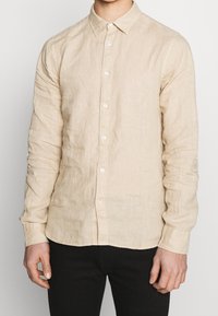 Camicia a maniche lunghe in lino beige con colletto button-down, caratterizzata da bottoni bianchi e dettagli di cucitura visibili lungo le maniche.