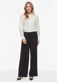 Balta palaidinė su vertikaliomis juostelėmis ir išsiuvinėtais akcentais, derinama su juodomis plačiomis kelnėmis ir juodais smailiais aukštakulniais.