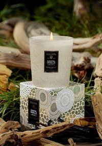 Kynttilä Voluspa Santal Vanille kirkkaassa lasissa, jossa on teksturoitu pinta; musta etiketti; sytytetty sydänlanka. Pakattu koristeelliseen laatikkoon, jossa on kukkakuosi.