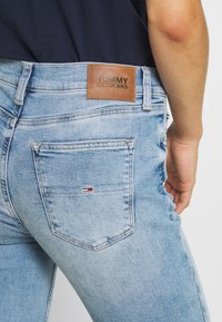 Vaqueros de denim azul desgastado con un parche de cuero marrón etiquetado "TOMMY JEANS" en la cintura, que incluye un bolsillo trasero con un pequeño detalle de logo.