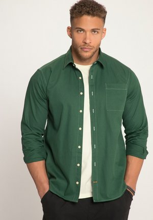 Green button-up shirt with a pocket, contrasting white stitching, and a relaxed fit. Worn over a light-colored t-shirt, paired with black pants.